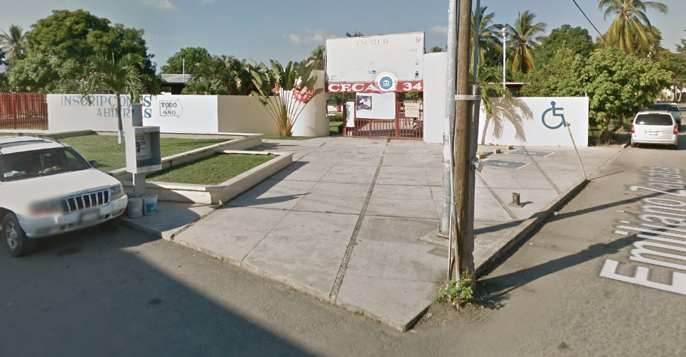 Fotografía en la que se muestra una explanada que tiene una jardinera. Al terminar la explanada hay una reja roja que permite el acceso a las instalaciones y encima de la reja dice CECATI 34.